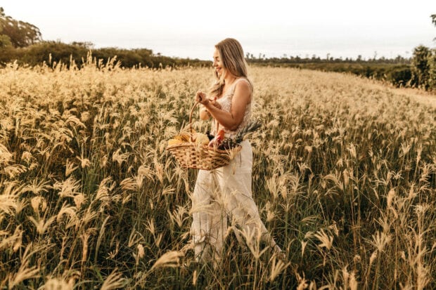 Woman walks through a golden field, carrying a woven basket filled with fresh produce and blooms