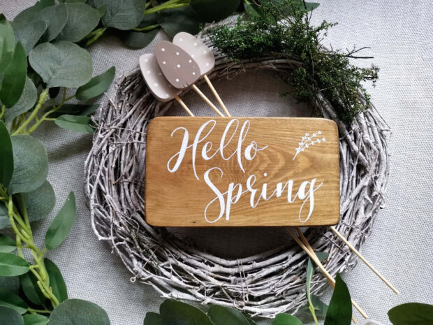 Rectangular wooden plaque displays white script text, resting on a rustic twig wreath, adorned with eucalyptus and decorative eggs