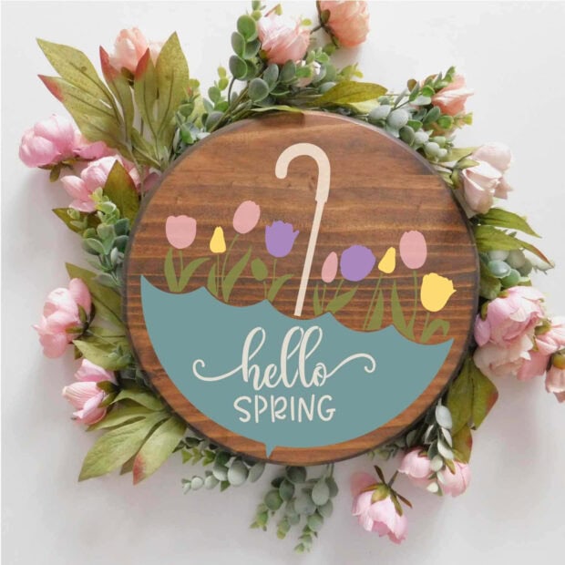 Round wooden sign showcases an umbrella design sheltering colorful tulips and white script text, encircled by pink and green faux flowers