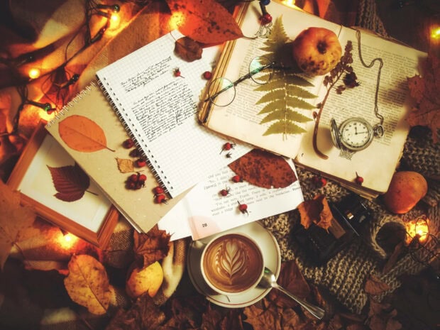 An aesthetic fall wallpaper showcasing a warm, cozy flat lay with coffee, an open book, scattered autumn leaves, and fairy lights, evoking a rich fall atmosphere