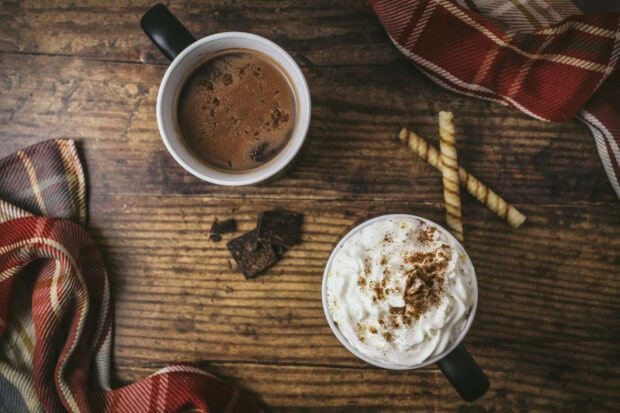 An aesthetic fall wallpaper displaying two cozy mugs of hot chocolate, one with whipped cream, on a wooden table with plaid fabric, creating a warm autumn scene