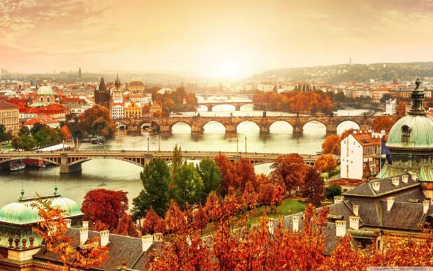 An aesthetic fall wallpaper showcasing the beautiful Prague cityscape with bridges and autumn trees under a warm sunset