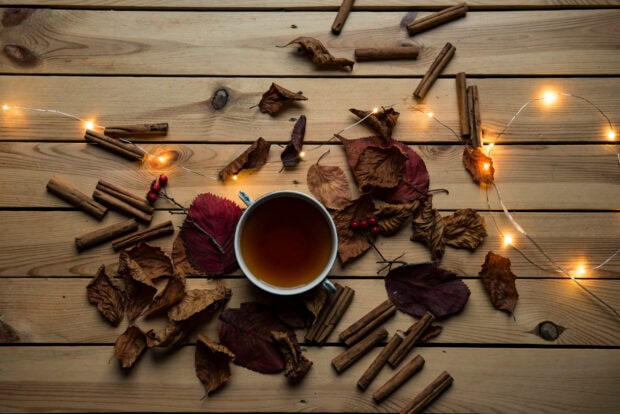 An aesthetic fall wallpaper featuring a cozy flat lay of a cup of tea, dried autumn leaves, cinnamon sticks, and warm fairy lights on a wooden surface