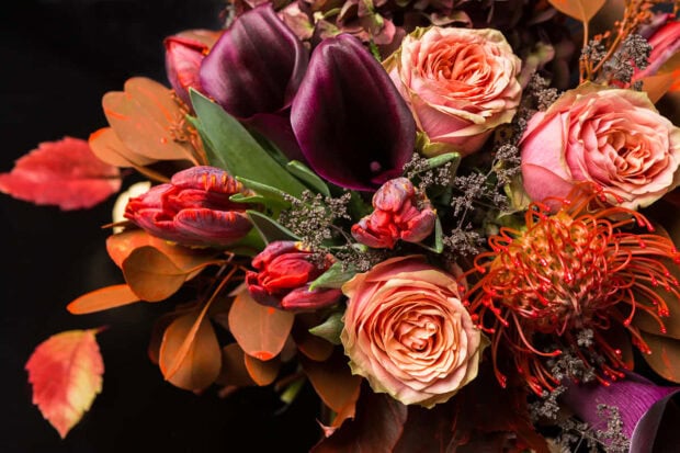 A beautiful Fall Flowers Wallpaper featuring a vibrant bouquet of roses, calla lilies, and autumn leaves against a dark background