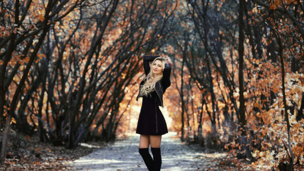 A charming Fall Season Wallpaper showcasing a blonde woman in a black dress standing on an autumn forest path