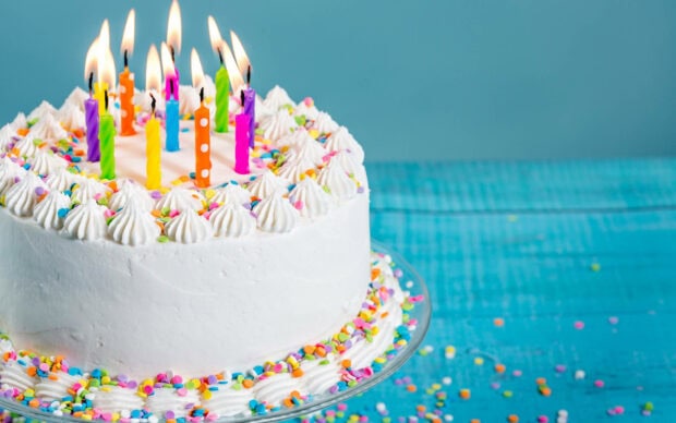 A cheerful birthday wallpaper featuring a white frosted cake with colorful sprinkles and lit candles on a blue wooden background
