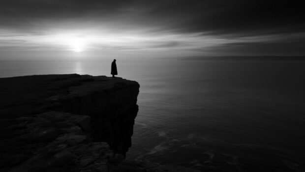 A dramatic black aesthetic wallpaper featuring a cloaked figure standing on a cliff edge overlooking a vast ocean at sunset
