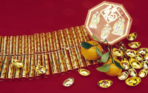 A festive Chinese New Year wallpaper displaying golden ingots, fresh oranges, and traditional firecracker decorations on a vivid red background