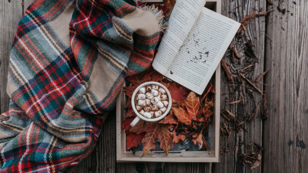 A cozy fall desktop wallpaper flat lay featuring a plaid blanket, hot chocolate with marshmallows, an open book, and autumn leaves on a wooden tray