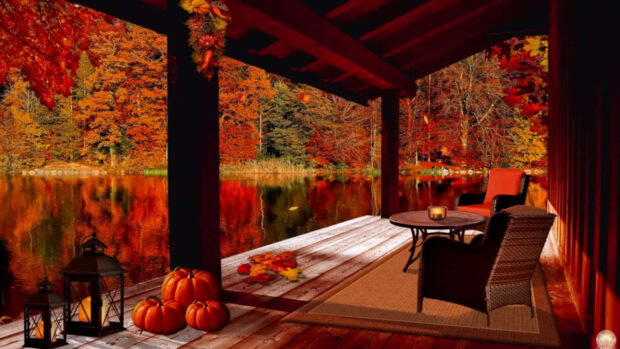 A cozy fall desktop wallpaper showing a wooden dock with pumpkins and seating overlooking a vibrant autumn lake with colorful trees