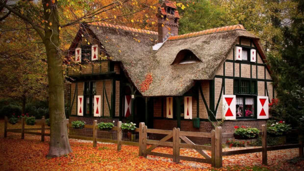 A charming cozy fall desktop wallpaper depicting a rustic cottage with a thatched roof and red shutters, surrounded by autumn leaves