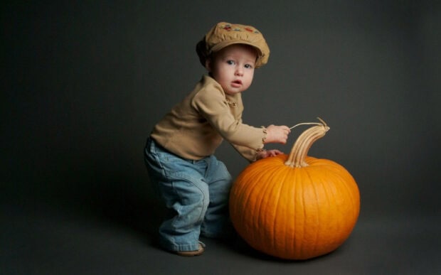 A cute Halloween wallpaper featuring a baby in a hat playing with a large orange pumpkin