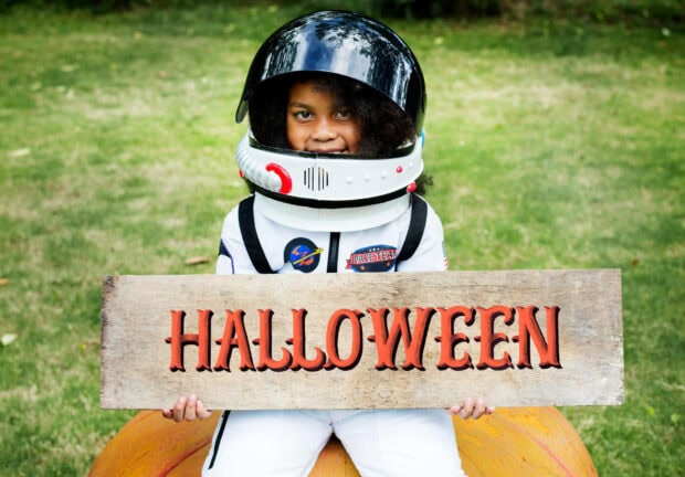 Cute Halloween wallpaper featuring a child in an astronaut costume holding a 'HALLOWEEN' sign on a pumpkin in a field