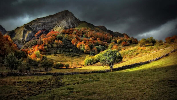 A beautiful Dark Fall Wallpaper featuring majestic dark mountains, vibrant autumn trees, and a green valley under a dramatic cloudy sky