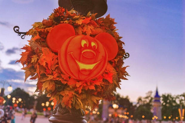 A vibrant Disney Halloween Wallpaper featuring a Mickey Mouse pumpkin decoration on a lamppost adorned with autumn leaves, set against a beautiful sunset sky with a distant castle