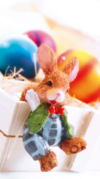 A delightful Easter Bunny Wallpaper featuring a cute ceramic bunny figurine in clothes with blurred colorful Easter eggs in the background