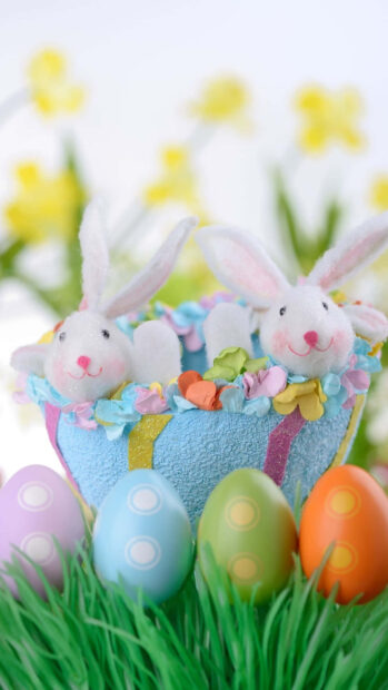 A vibrant Easter Bunny Wallpaper featuring two white bunny figurines in a decorative blue basket surrounded by colorful Easter eggs on green grass with a soft, blurred background