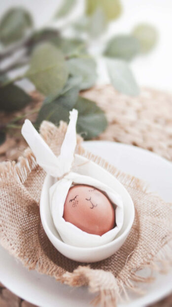 Easter Egg Wallpaper showing a cute egg with a sleeping face drawing, nestled in a bowl on a burlap mat