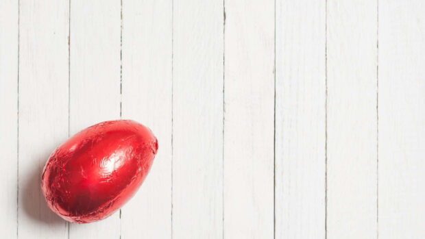 A minimalist Easter Egg Wallpaper showcasing a single red foil-wrapped chocolate egg on a white wooden plank background