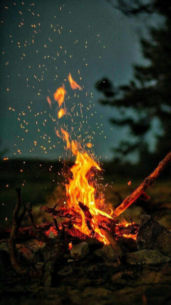 A cozy fall aesthetic iPhone wallpaper featuring a crackling bonfire with flying sparks against a dark night sky