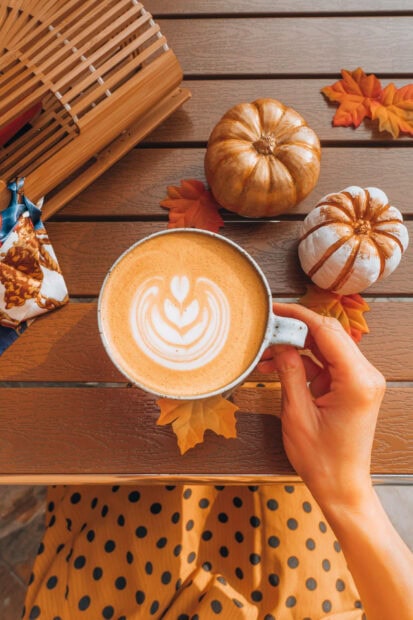 A cozy Fall Aesthetic Iphone Wallpaper featuring a hand holding a latte with art, surrounded by small pumpkins and autumn leaves on a wooden table