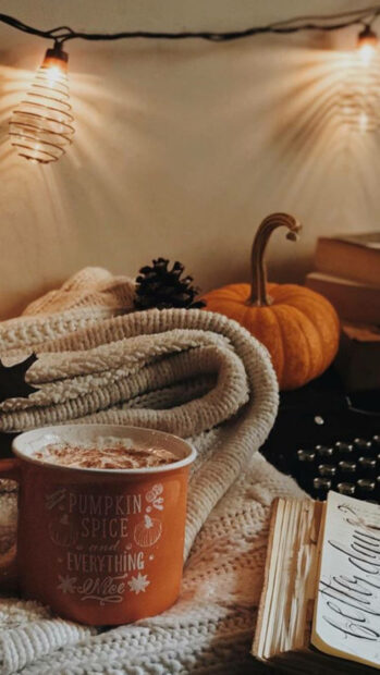 A cozy fall season wallpaper featuring a "Pumpkin Spice and Everything Nice" mug, a small pumpkin, a pinecone, and warm string lights in an indoor setting