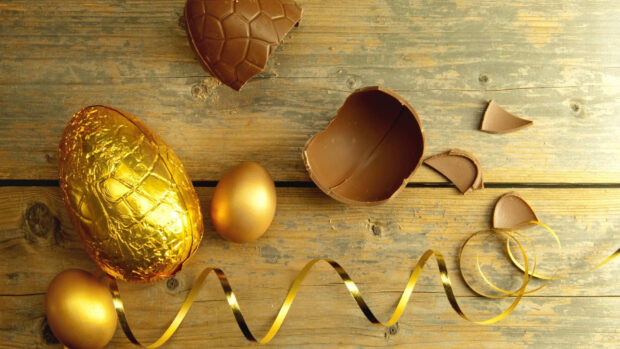 A top-down view of golden and broken chocolate Easter egg wallpaper with a ribbon on a rustic wooden table