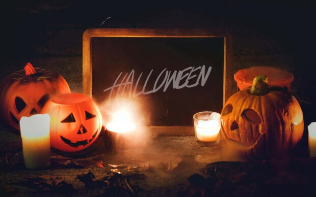 A dark scene with "HALLOWEEN" on a blackboard, surrounded by glowing candles and pumpkins, makes a cozy Halloween Macbook wallpaper
