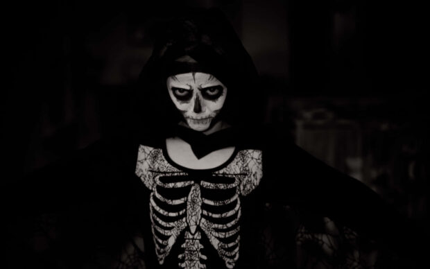 A spooky Halloween Skeleton Wallpaper featuring a child in a skeleton costume with face paint and a dark hood, set against a dark background