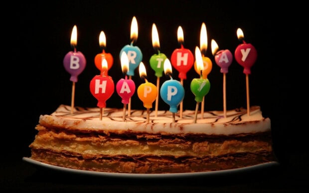 Its My Birthday Wallpaper showing a delicious layered cake with colorful burning candles spelling 'Happy Birthday' against a dark background