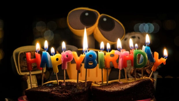 Its My Birthday Wallpaper showcasing a chocolate cake with colorful 'Happy Birthday' letter candles and blurred Wall-E eyes in the background