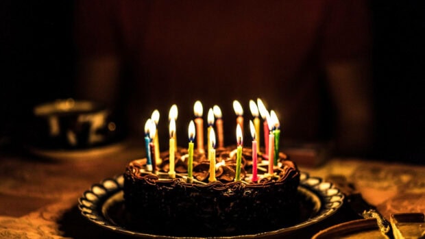 Its My Birthday Wallpaper showing a chocolate birthday cake with numerous lit colorful candles in a warm, dimly lit setting