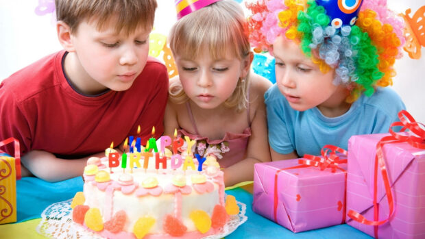 Its My Birthday Wallpaper showing three kids, one in a colorful wig, blowing out candles on a festive birthday cake with gifts nearby