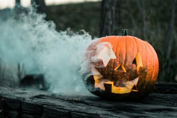 Kawaii Halloween wallpaper featuring a spooky carved pumpkin emitting smoke on a dark wooden log