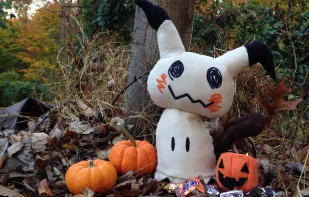 An adorable Pokemon Halloween Wallpaper featuring a Mimikyu plush toy sitting among autumn leaves, small pumpkins, and candy in a natural outdoor setting