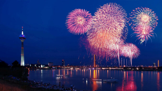 A stunning New Years Eve Wallpaper featuring vibrant pink fireworks exploding over a city river with a prominent tower and illuminated skyline at night