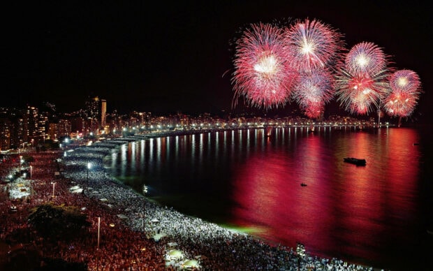 A vibrant New Years Eve Wallpaper showcasing a crowded beach and city skyline illuminated by red fireworks reflecting on the water