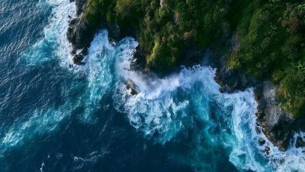 An aerial Ocean Wallpaper featuring powerful waves crashing against a rugged rocky coastline covered with lush green trees, showcasing stunning blue and turquoise waters