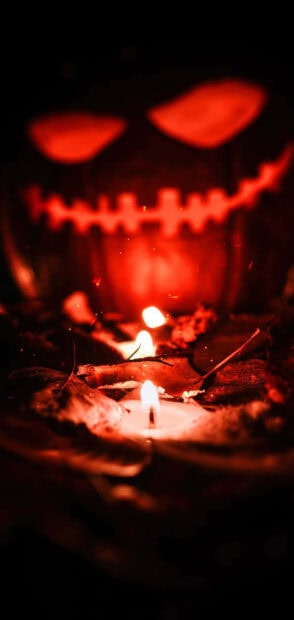 A dark preppy Halloween wallpaper featuring a glowing red jack-o'-lantern face with lit candles among autumn leaves