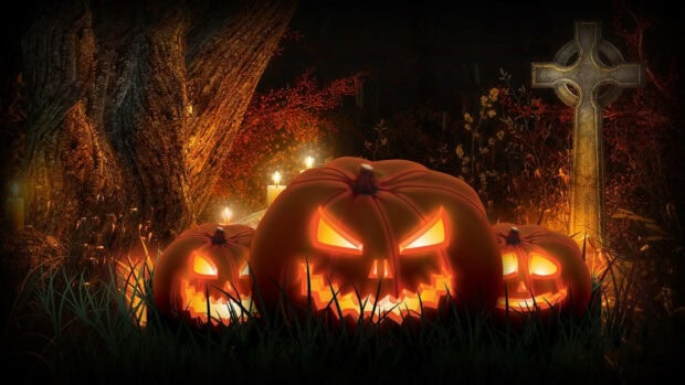 A spooky Halloween iPad wallpaper showing three glowing jack-o'-lanterns in a dark graveyard next to a Celtic cross