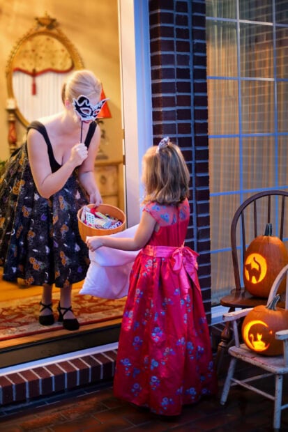 A Cute Halloween Wallpaper showing a little girl trick-or-treating from a woman in a cat mask, with carved pumpkins on the porch