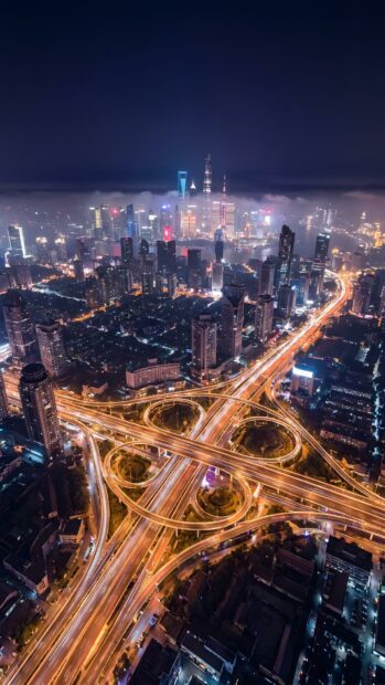 An breathtaking aerial City Wallpaper showcasing a bustling city at night, with illuminated highways forming intricate patterns and towering skyscrapers piercing through the misty sky