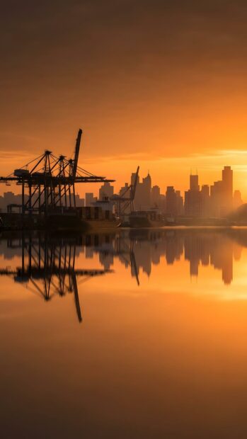 A serene City Wallpaper capturing a city skyline with port cranes silhouetted against a vibrant orange sunset, reflecting beautifully on the still water