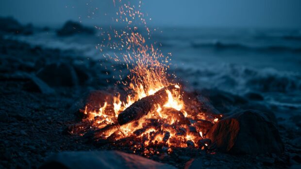 A serene fire wallpaper of a campfire burning on a rocky beach with ocean waves in the background at night