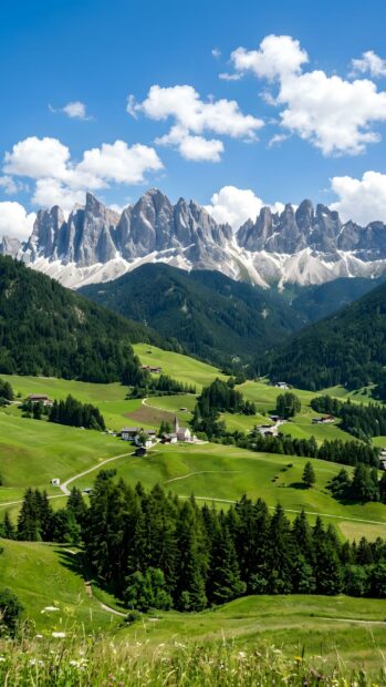 A picturesque landscape wallpaper showing a green valley with a small village and pine forests nestled below dramatic jagged mountains under a clear blue sky