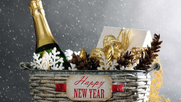 A cozy New Year Aesthetic Wallpaper showing a basket with a champagne bottle, gift, pinecones, and 'Happy New Year' tag amidst falling snowflakes