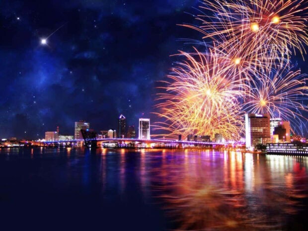 A stunning New Year Aesthetic Wallpaper featuring a city skyline at night with vibrant fireworks exploding over the water and a shooting star in the dark sky