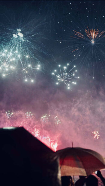 New Year Aesthetic Wallpaper showing colorful fireworks exploding in a cloudy night sky with silhouettes of people holding an umbrella