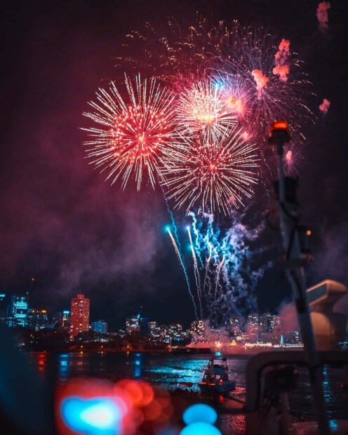 A vibrant New Year Aesthetic Wallpaper featuring colorful fireworks exploding over a city skyline at night, viewed from a boat