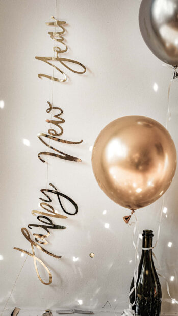A festive New Year Aesthetic Wallpaper showing a gold "Happy New Year" banner, shiny gold and silver balloons, and a champagne bottle against a bright background